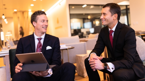 Two men in suits sit together smiling while one holds an iPad.