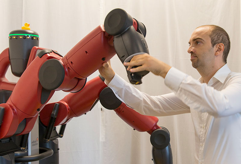 A man manually manipulated a robot's arm. The caption reads: Heni Ben Amor works with a Baxter robot in his lab. Photo by Jessica Hochreiter.