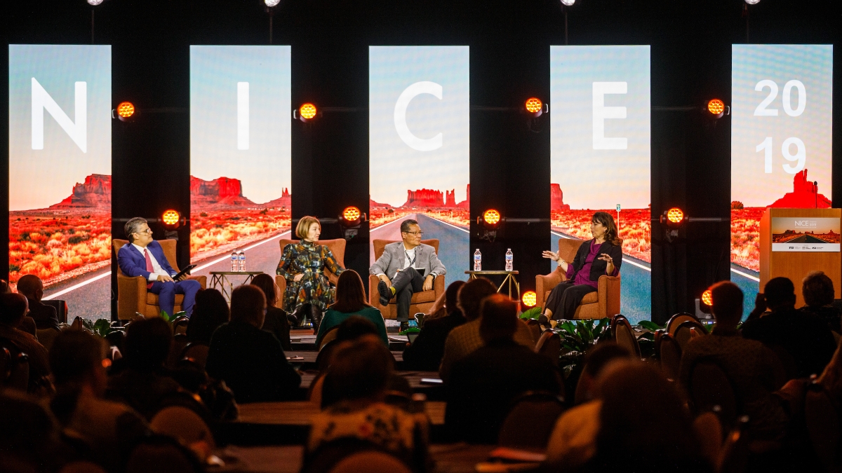 Four people sit conversing on a stage with "NICE 2019" displayed behind them. 
