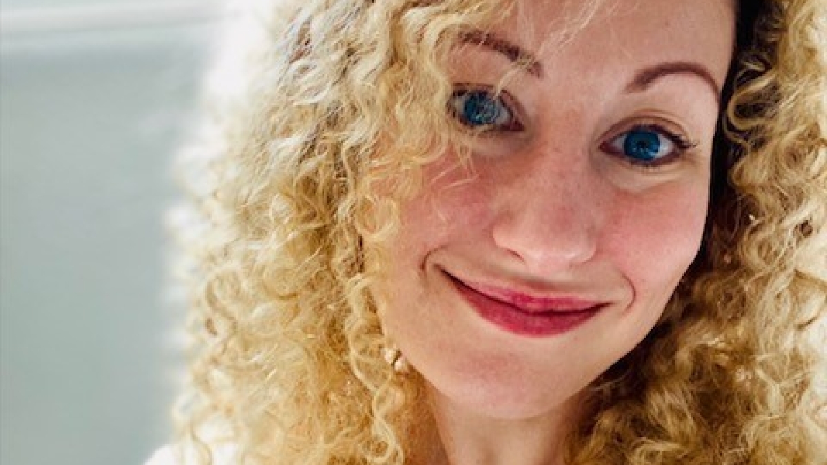 A woman named Katie Guerin has curly blonde hair and wears a flowered shirt as she poses for a selfie.