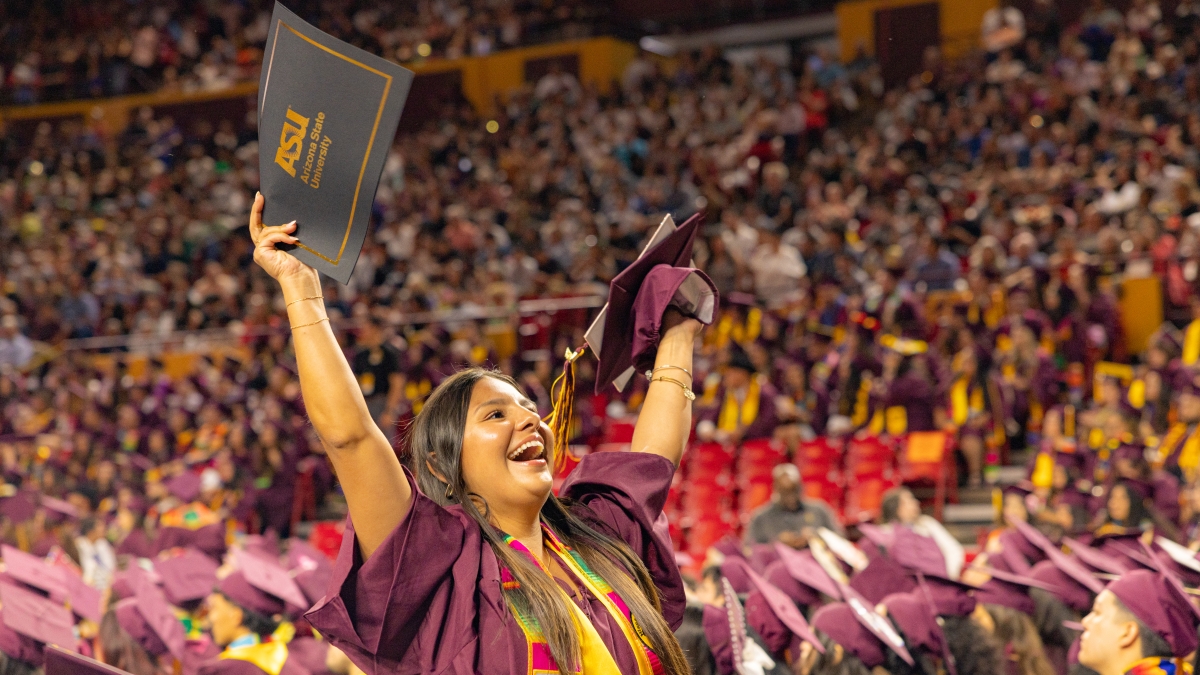 Student in graduation gown standing in a packed stadium full of graduates, holding up a graduation cap and ASU-branded certificate