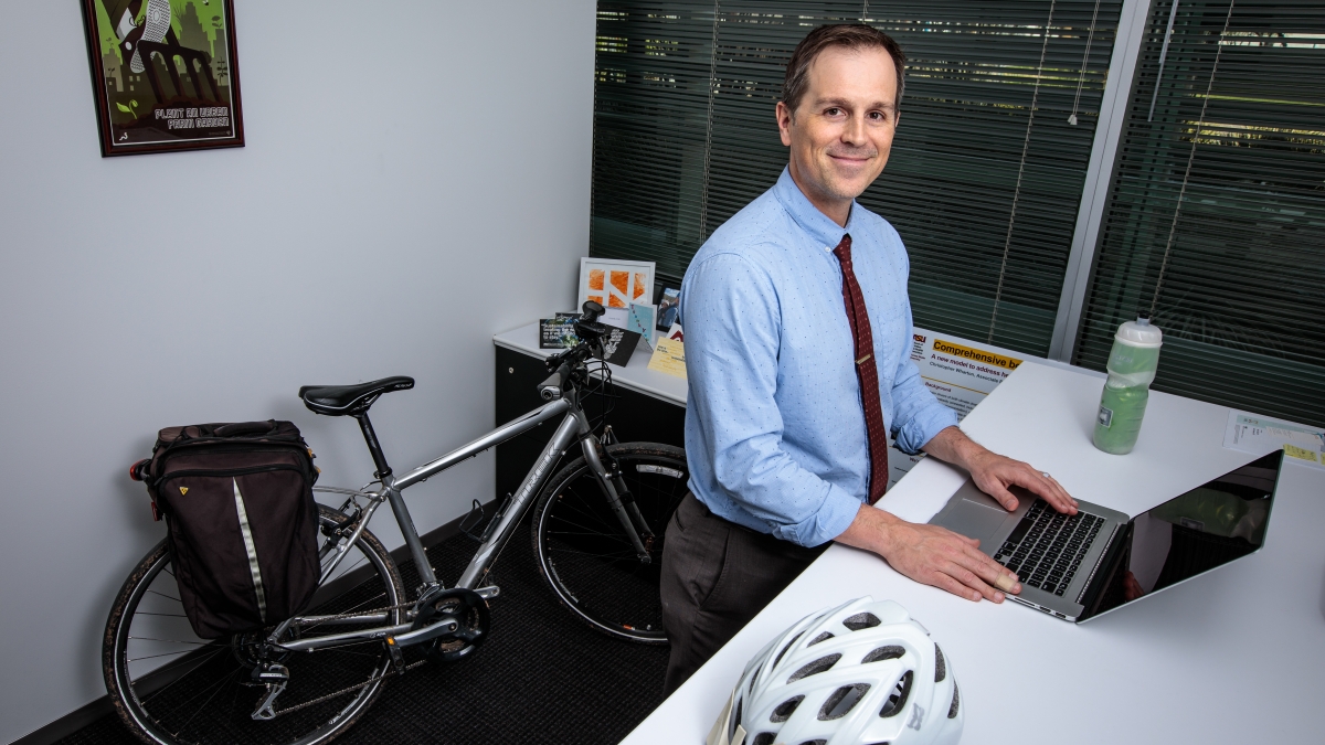 man standing at desk with hands on laptop and a bike leaning on the wall behind him