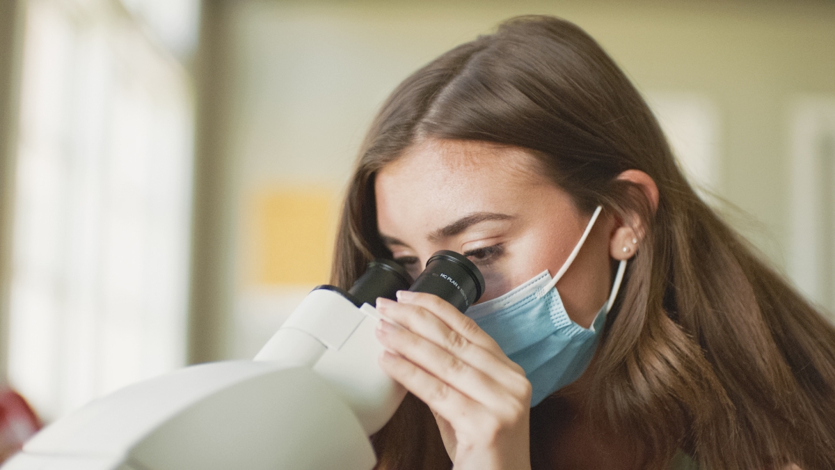 A student wearing a mask looks into a microscope