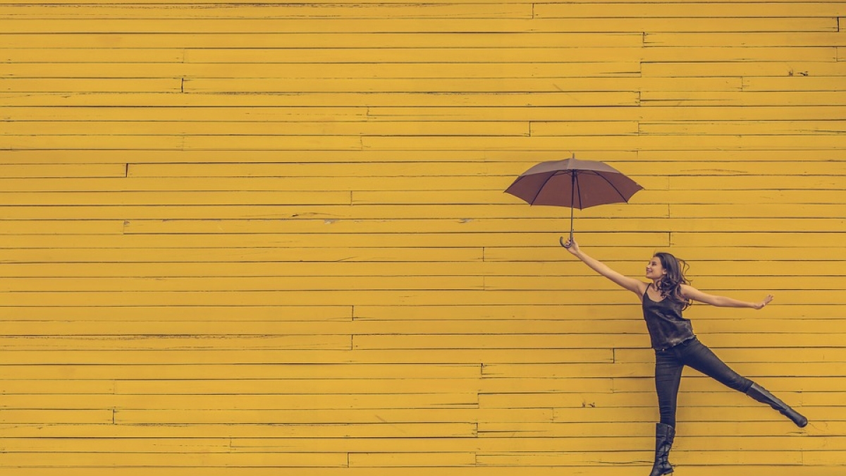 woman smiling and jumping while holding an umbrella in front of a yellow brick wall