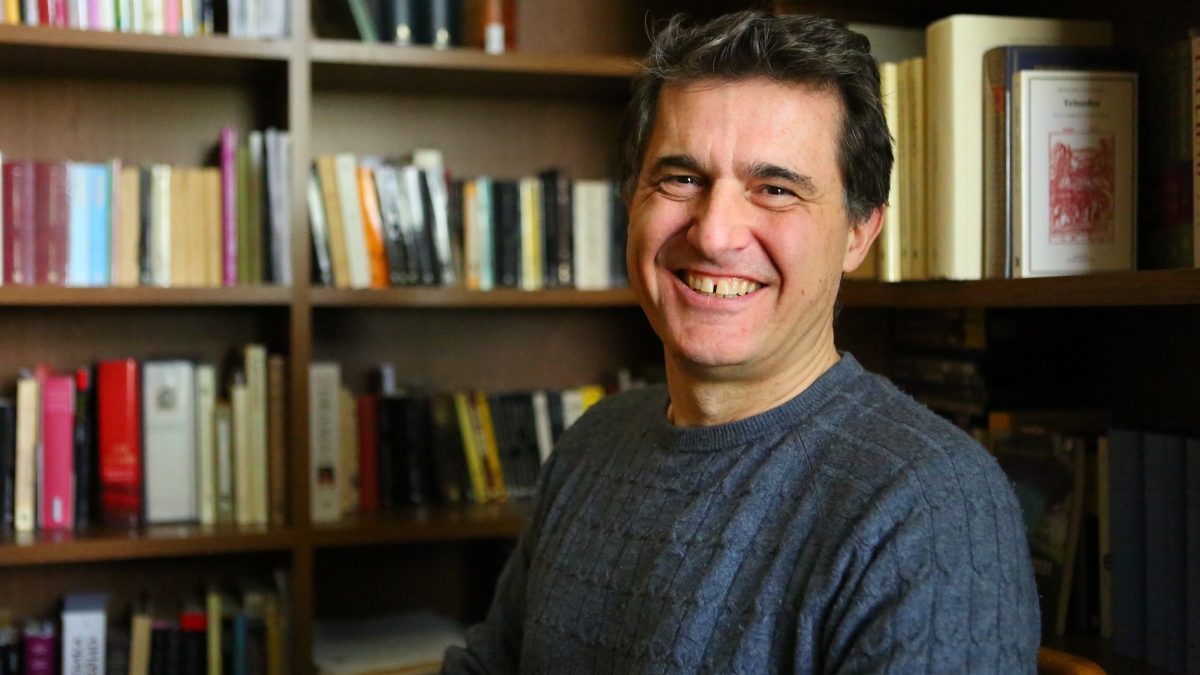 Professor Juan Pablo Gil-Osle sits in his office on ASU's Tempe campus. The top half of his body is visible, he is wearing a navy sweater, has short brown hair, and is smiling. Behind him are brown bookshelves filled with a variety of books.