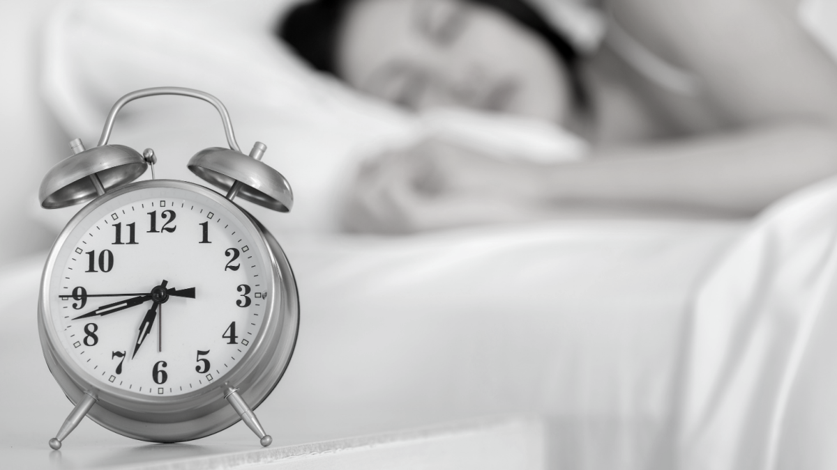 Alarm clock in the foreground with woman sleeping in the background