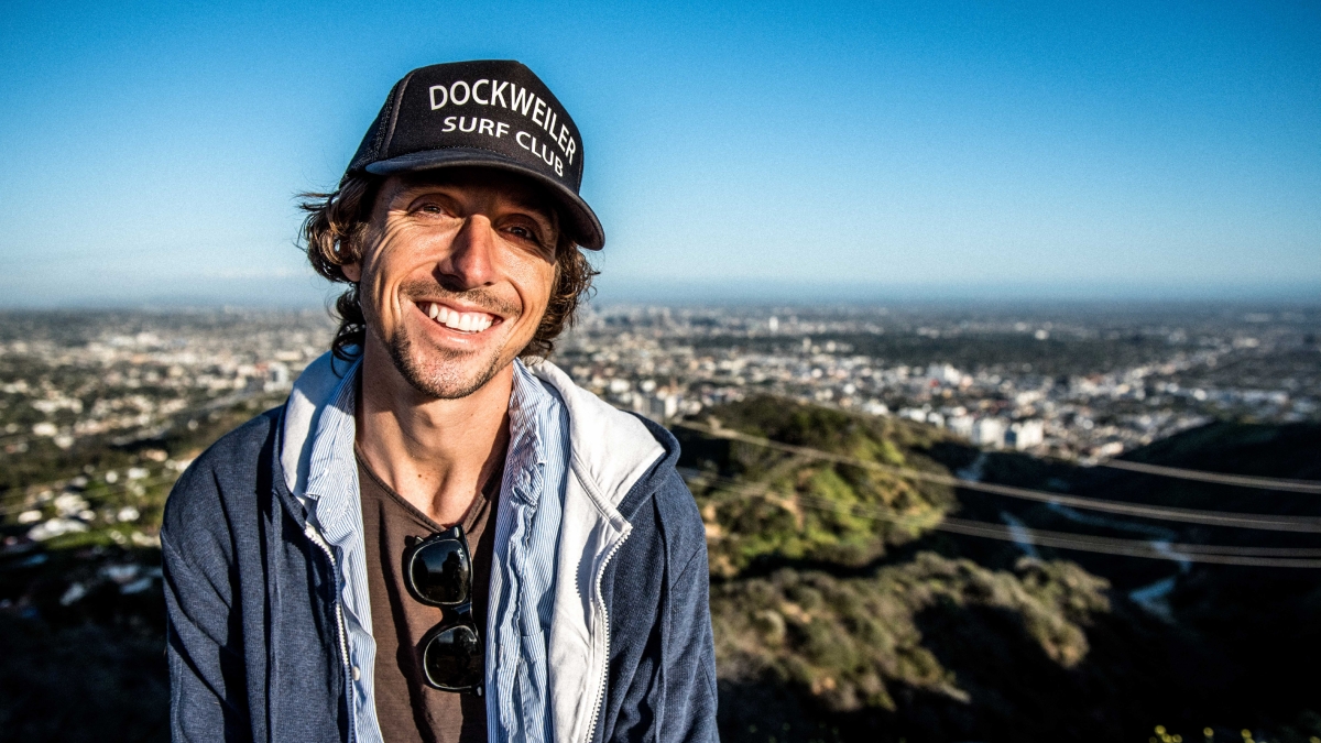 Author Nic Sheff smiles atop a mountain in a black baseball cap reading "Dockweiler Surf Club" and blue open jacket