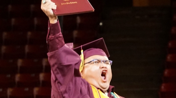 Graduate wearing robe, cap and stole holding up a diploma with a victorious expression.