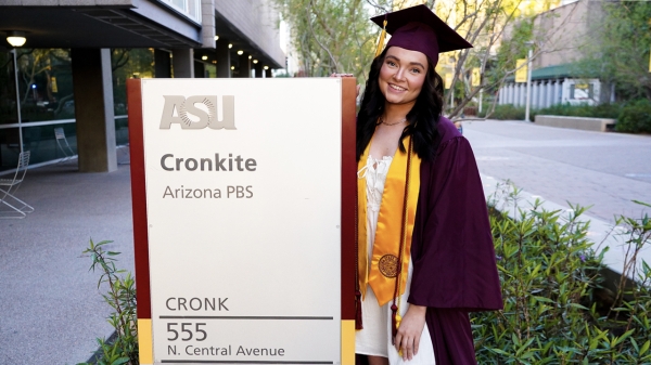 Rebecca Striffler standing next to a Cronkite building sign dressed in grad gown, cap and stole.