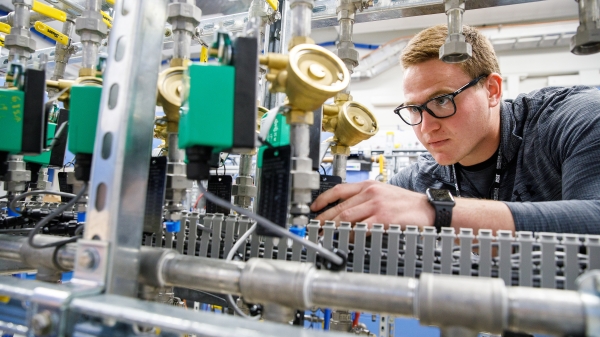A young man in glasses examines a piece of scientific equipment. The caption reads: Alex Gardeck, a mechanical engineering student, examines one of the precision thermal trim unit water systems that is used to control the temperature of various components