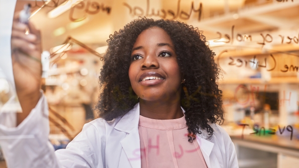 Woman with dark skin and hair wearing a white lab coat and writing equations on a board.