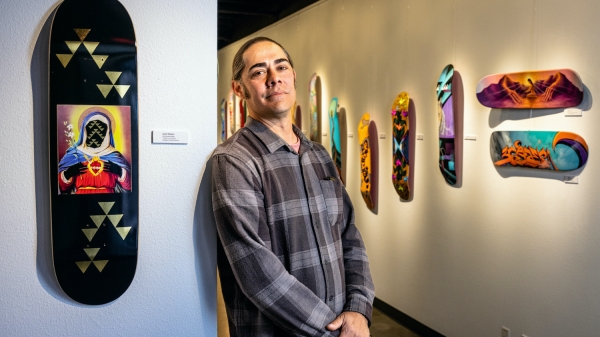 ASU Assistant Professor Jacob Meders stands in front of a skate board he painted, called "Creator is Woman" at ASU West ArtSpaceWest.