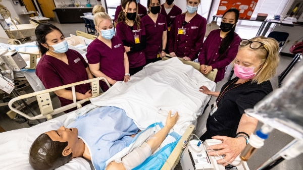 Nursing students look on as an instructor demonstrates at an IV pump next to a manikin. The students are wearing maroon scrubs and surgical masks 