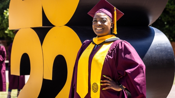 A woman stands proudly in graduation cap and gown in front of a giant 2022 sign