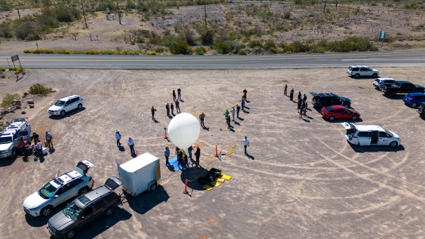 View from above of a group of people and cars in a desert area prepping a large white balloon.