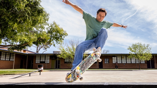 Maurice Crandall on a skateboard mid-jump, in an outdoor setting, wearing jeans and a green shirt.