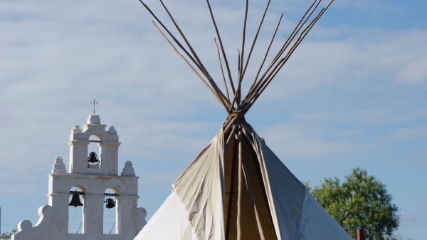 The top of a teepee is seen in the foreground with a Spanish mission in the background