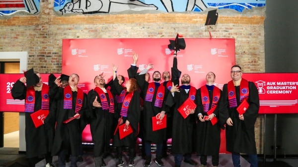 People wearing black graduation robes and red stoles throw their caps into the air in celebration.