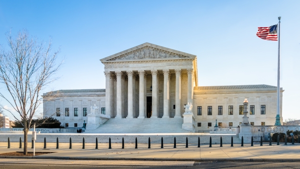The U.S. Supreme Court building stands under a blue sky with the American flag waving.