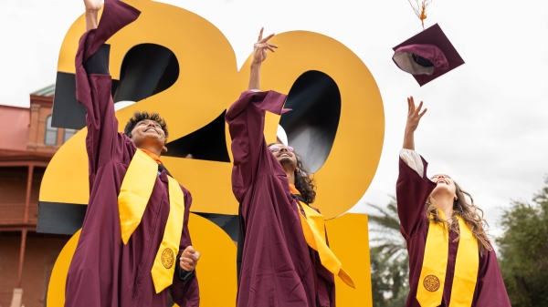 Three ASU grads in cap and gown throw their caps into the air in front of a giant 2024 sign