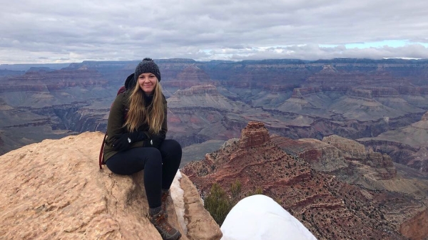 Sierra Boyd sits on a boulder in the foreground with a sprawling vista of red canyons and cloudy skies in the background