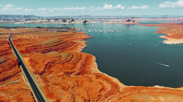A large blue desert reservoir shines amongst bright red and peach toned sandstone rock formations with a long road leading to a marina.