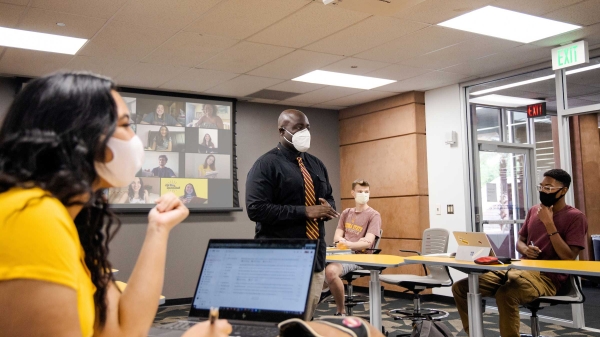 A teacher wearing a mask speaks to a socially distanced classroom of masked college students with a Zoom screen behind him of more students