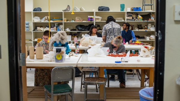 Students creating artwork while sitting at tables, sculptures lined on shelves behind them.