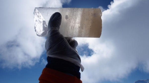 Gloved hand holding an ice core from Antarctica up to a cloud-filled blue sky.