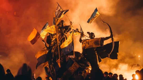 Statues are adorned with Ukraine flags as smoke fills an evening sky around them