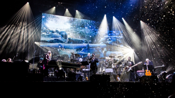 The ensemble of Mannheim Steamroller Christmas performs onstage with an image of snowy mountains behind them and lights imitating snowfall.