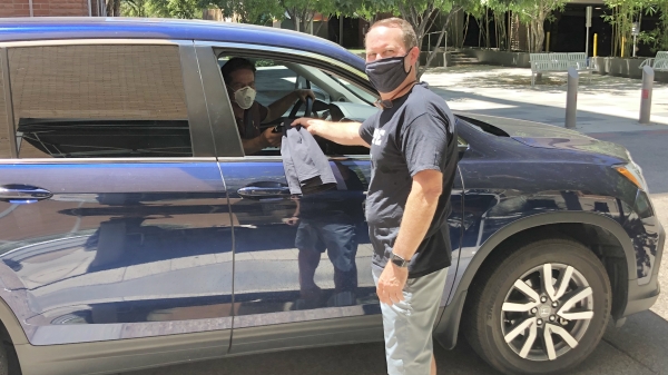 ASU Enterprise Partners CEO Dan Dillon wears shorts, a black t-shirt and black mask and passes out a black t-shirt to employee Peter Means through the passenger window of his blue vehicle. The employee wears a white face mask. 