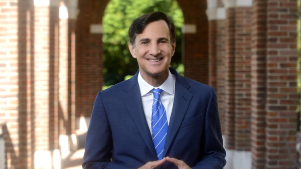Ronald J. Daniels, president of Johns Hopkins University, is pictured in an outdoor corridor, smiling at the camera with his hands together.