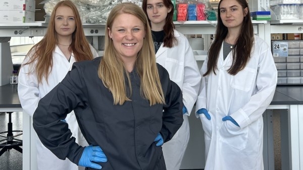 Candace Lewis and her lab assistants, standing behind her, posing for a photo in lab gear and in a lab setting.