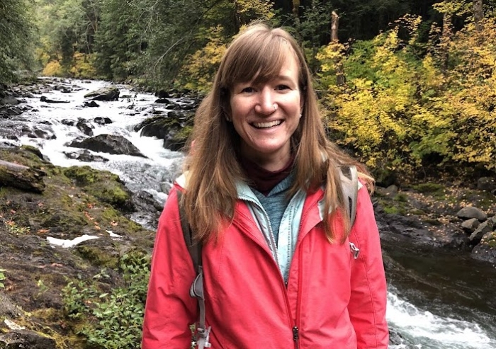 A woman in a red jacket smiling in front of a river.