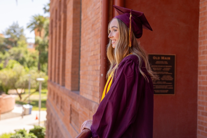 Aubrey Lines dressed in ASU graduation gear standing in front of Old Main looking out and smiling
