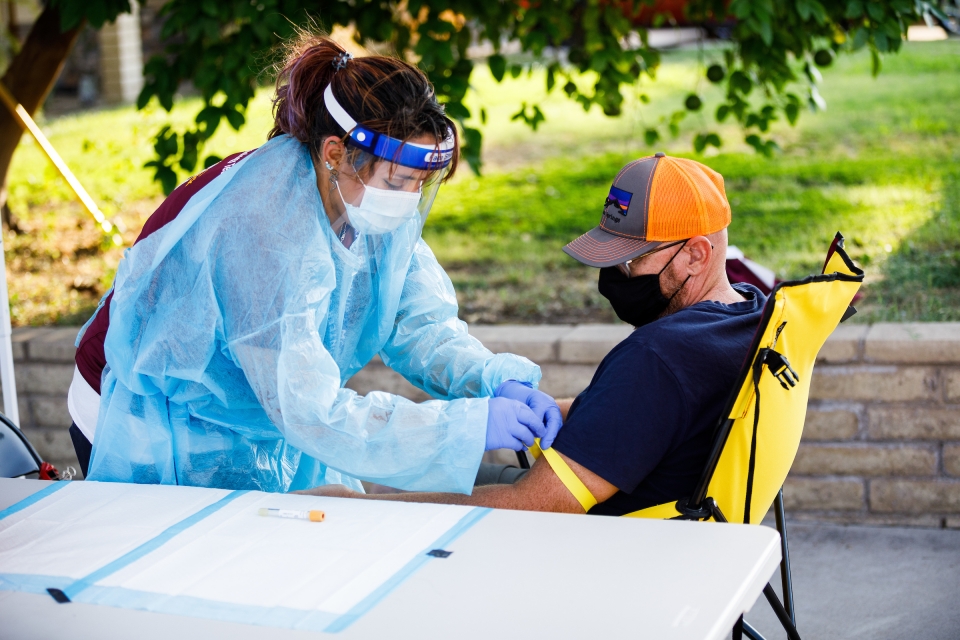 A woman in full personal protective gear indicates the site of blood draw on a man's arm.