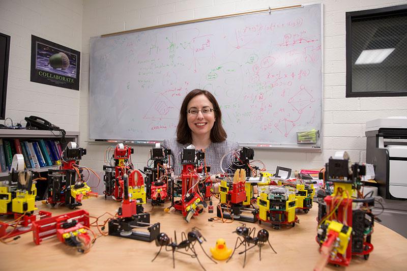 A woman smiles, surrounded by small robots. 