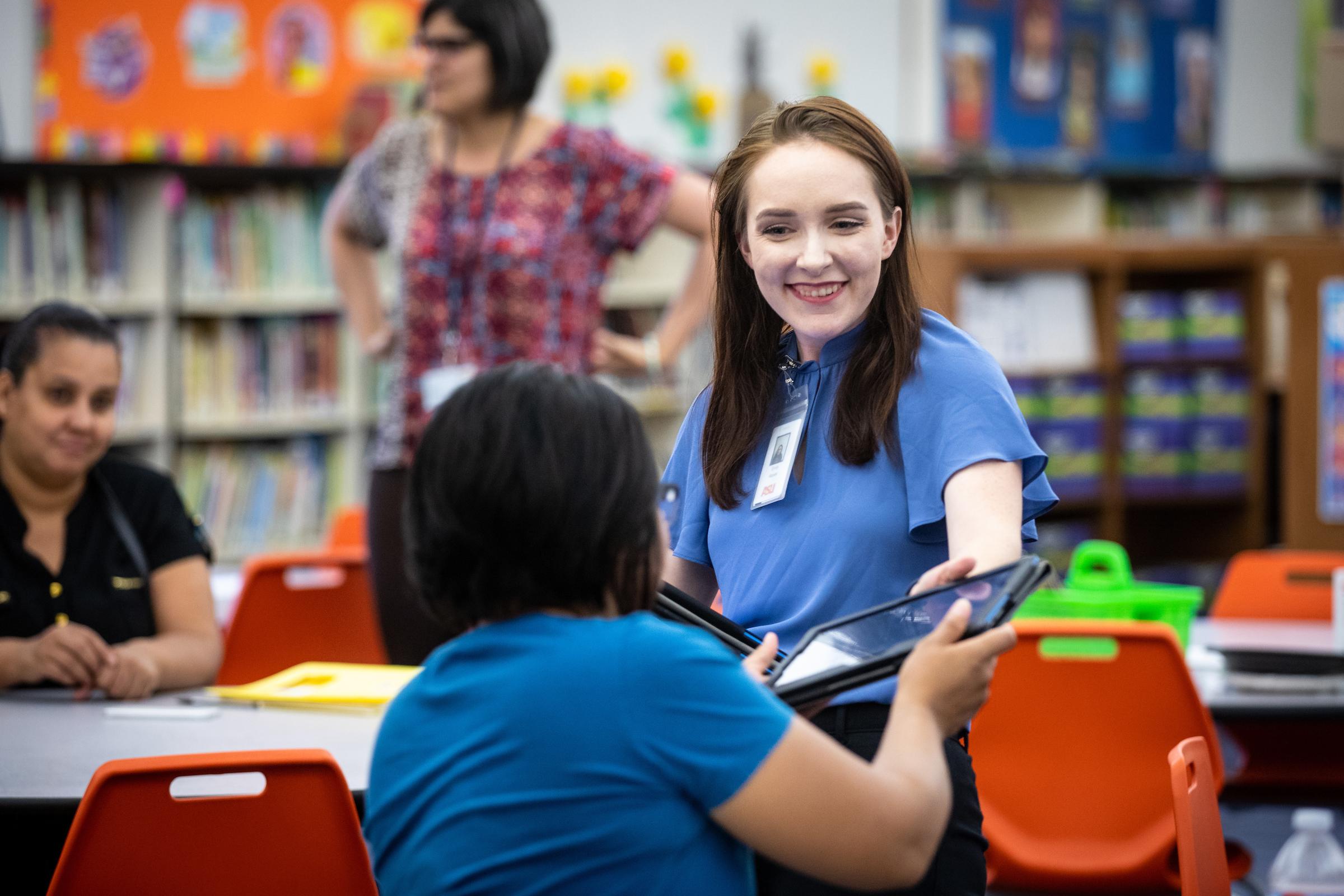 woman smiling as she hands a tablet to another woman in a library setting